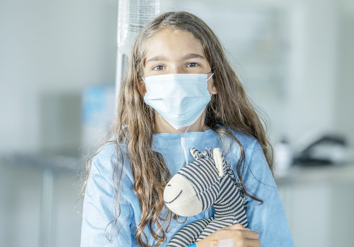 10 year old girl sitting in hospital wearing a protective face mask holding her favourite stuffed animal also wearing a protective face mask during COVID-19.